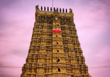 A towering golden gopuram (temple tower) against a vibrant purple and pink twilight sky. The intricate carvings and details of the gopuram are illuminated, and a small red banner or flag is visible near the top.