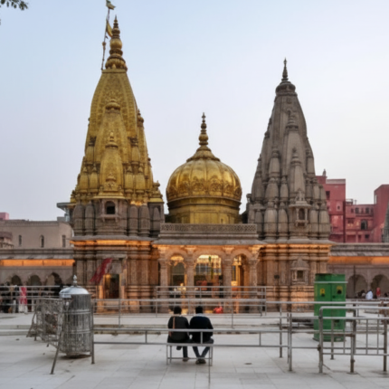 The magnificent Kashi Vishwanath Temple complex in Varanasi at dusk, showcasing its iconic golden spires and domes, surrounded by traditional Hindu architecture and a few devotees in the courtyard.