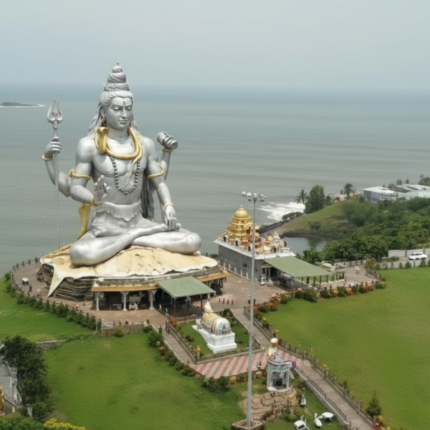 An aerial view of the colossal silver-colored statue of Lord Shiva in a meditative pose, located at Murudeshwar, Karnataka, overlooking the Arabian Sea under an overcast sky. The statue is adorned with golden accents and surrounded by green landscape and temple structures.