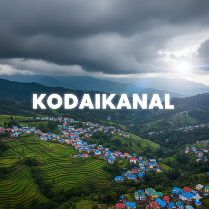 An aerial view of a picturesque hill station village in Kodaikanal, with brightly colored houses nestled among lush green terraced farmlands under a dramatic cloudy sky, with "KODAIKANAL" text overlay.
