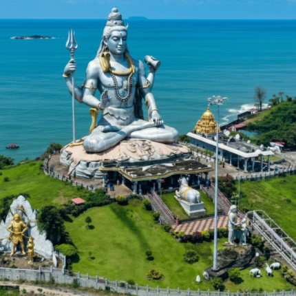 An impressive aerial view of the colossal silver Lord Shiva statue in Murudeshwar, surrounded by lush green hills, overlooking the vast blue Arabian Sea, with a temple complex and other smaller statues in the foreground.